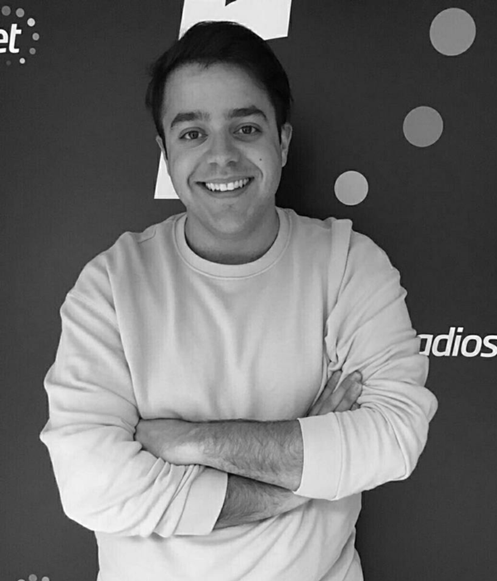 Imagen en blanco y negro de David Insua. Posa ante un cartel. Está sonriente y lleva una sudadera remangada. Está con los brazos cruzados.