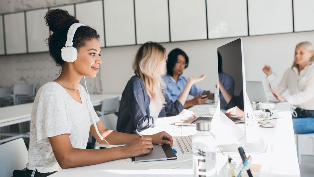joyful-african-web-developer-working-on-new-project-while-listening-music-in-white-headphones-black-female-designer-doing-her-job-in-office-with-talking-colleagues