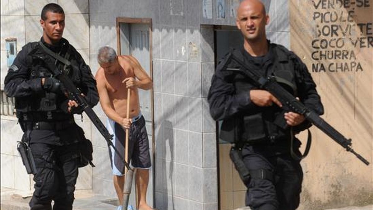 Dos policías patrullan una calle de la favela Grota mientras un hombre asea el frente de una casa este 29 de noviembre un día después de que las fuerzas de seguridad tomaran varias barriadas en Río de Janeiro (Brasil). EFE