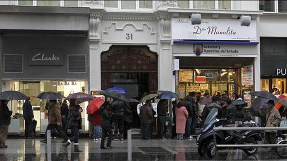 Decenas de personas que esperan su turno para comprar lotería en una administración en la Gran Vía de Madrid se cubren de la lluvia con un paraguas por el temporal atlántico que afecta a la Península Ibérica, caracterizado por una masa de aire "húmeda y cálida", que comenzará a remitir a partir de mañana. EFE