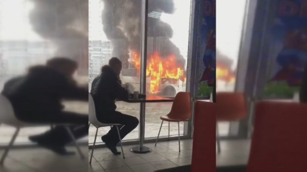 Un joven disfruta impasible de un café a escasos metros de un autobús en llamas