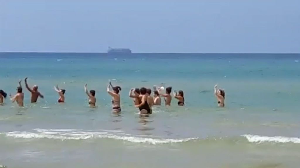 Turistas bailando en la playa y un barco repleto de inmigrantes al fondo en la Italia de Salvini