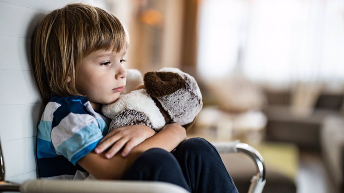 Depressed little boy embracing his teddy bear while relaxing on a chair during home quarantine.