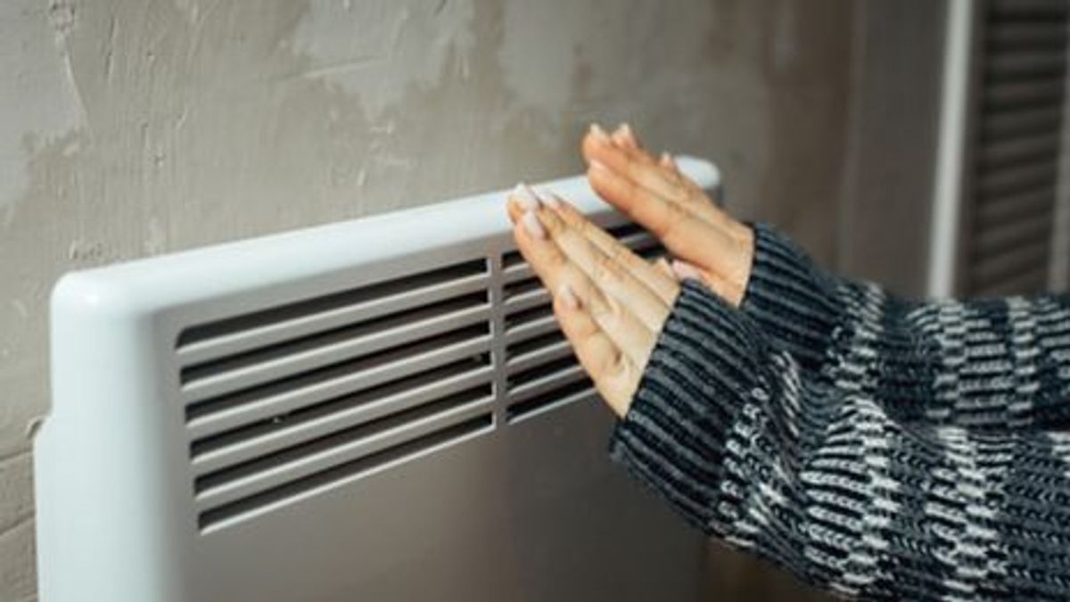 a woman warms her hands at the radiator in a cold house, problems with heating, heating the room with an electric convector a woman warms her hands at the radiator in a cold house, problems with heating, heating the room with an electric convector