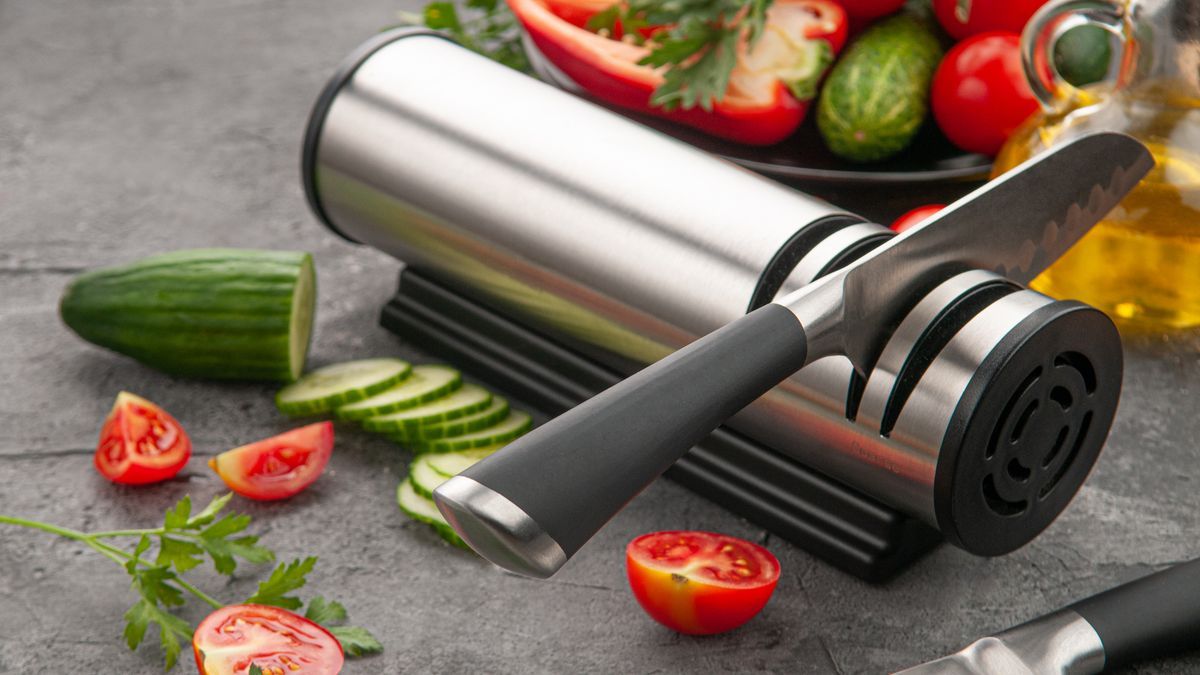 Electric knife sharpener. Cylindrical shape made of metal and plastic. A gadget for the kitchen and interior. Gray concrete background. Nearby are kitchen knives and salad ingredients.