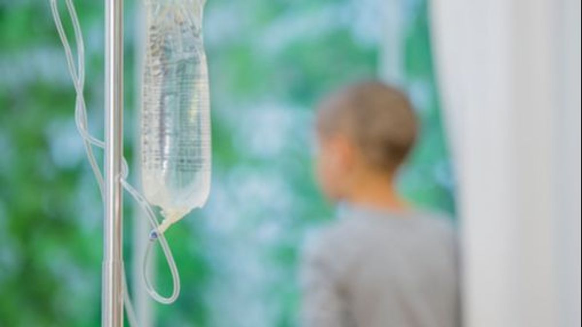 A little boy with a shaved head is looking out of the window with chemotherapy in the foreground. He is battling cancer. A little boy with a shaved head is looking out of the window with chemotherapy in the foreground. He is battling cancer.