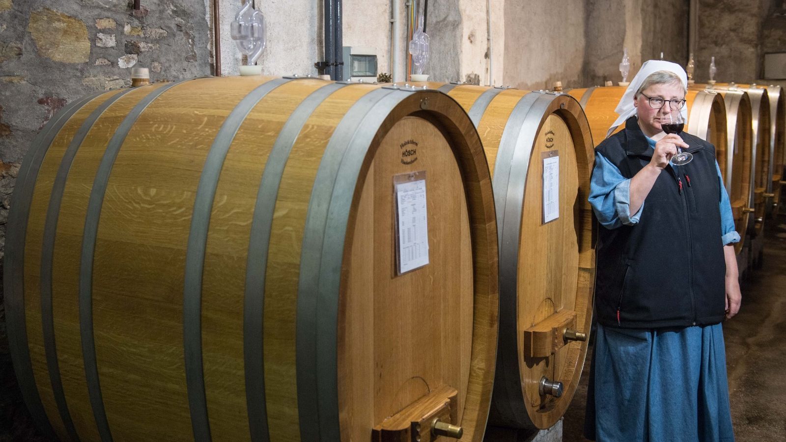 Una monja cata una copa de vino junto a varias botas