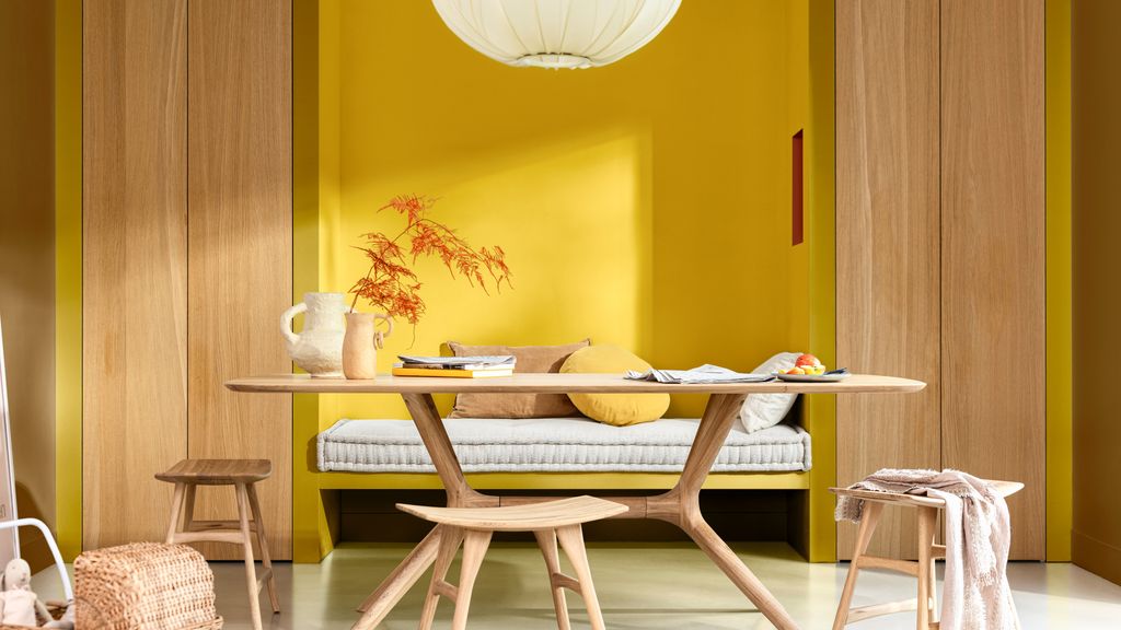 Comedor con mesa y banco de madera, pared amarlla y lámpara de techo blanca