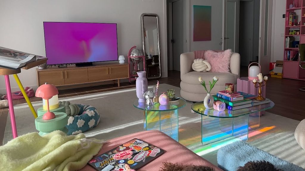 Salón Laura Escanes con librería rosa, mueble de televisión de madera, mesas auxiliares de cristal, butaca blanca, alfombra en color crudo y jarrones y lámparas de colores