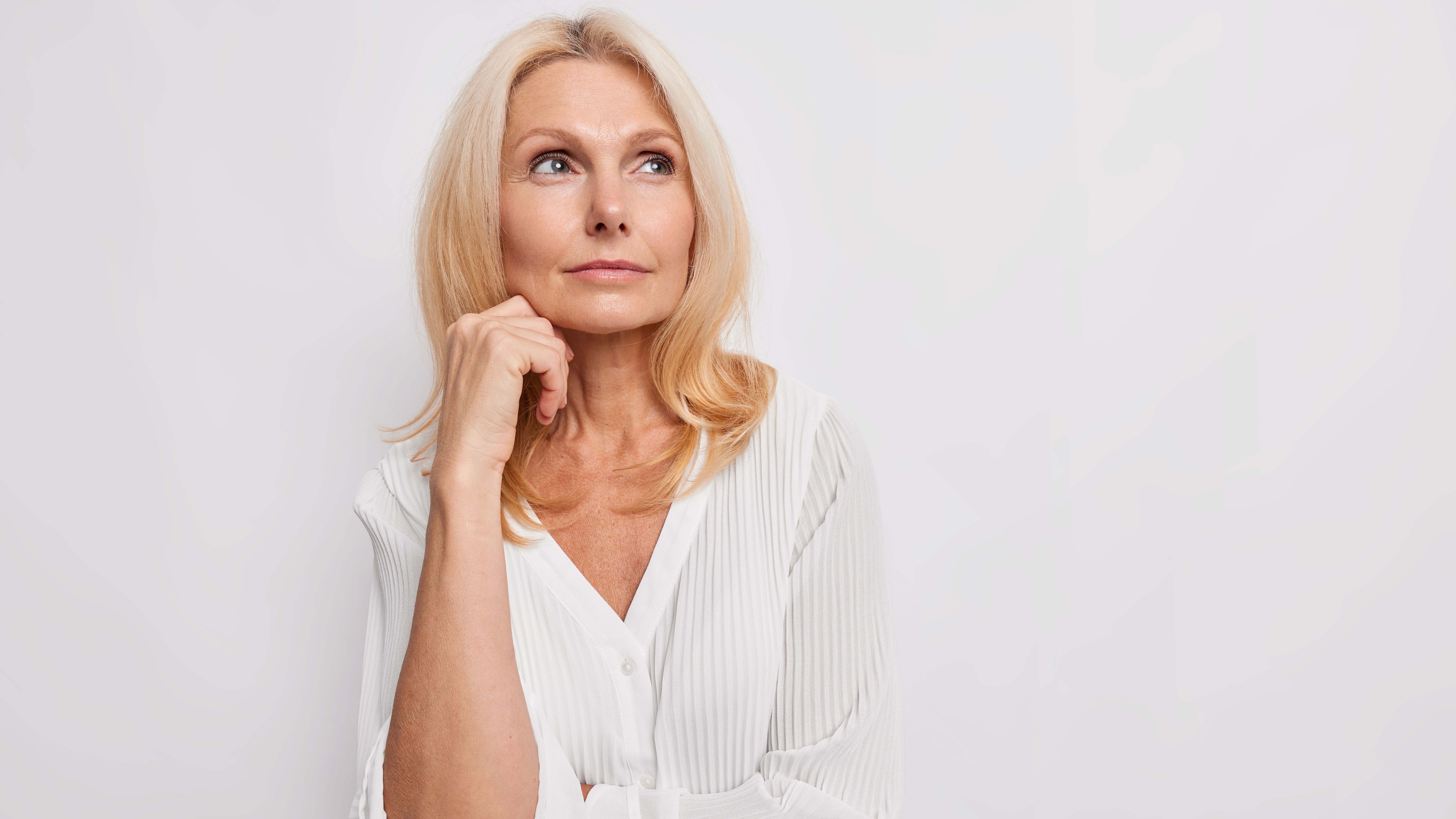 Thoughtful blonde middle aged woman ponders on something keeps hand near face has healthy skin minimal makeup makes choice wears white blouse poses indoor blank copy space for your promotion