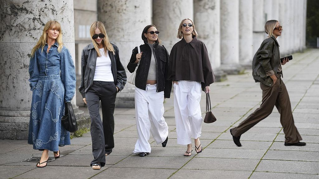 MUNICH, GERMANY - SEPTEMBER 2: Anna Winter (left) is seen wearing ultra wide-leg white denim pants from The Frankie Shop; a cropped brown-grey ribbed knit turtleneck top from Zara; an oversized black suede jacket with flap pockets and zip closure from Zara; a pair of square-toe black leather Pierced Mules with a silver-tone toe ring detail from Tory Burch; a light cream colored small Romy hobo leather bag with slim shoulder strap from Tory Burch; rectangular sunglasses with burgundy acetate frames and dark blue shaded lenses from Ray-Ban; her brown hair is worn sleek and straight with a center part; Marlies Pia Pfeifhofer (right) is seen wearing a voluminous dark brown cropped trench coat cape with wide draped sleeves from COS; white barrel trousers with ankle length and wide leg silhouette from COS; a pair of black flip-flop heeled sandals with square toes from H&M; a dark brown suede The New York shoulder bag with top handles, triangular fold design and golden hardware from DeMellier; round sunglasses with slim golden frames and dark-shaded lenses from Persol; her blonde bob hair is styled in a sleek, chin-length cut with a center parting on September 2, 2025 in Munich, Germany. (Photo by Moritz Scholz/Getty Images)