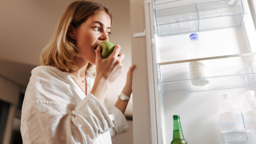  Mujer joven comiéndose una manzana mientras mira lo que hay en el frigorífico