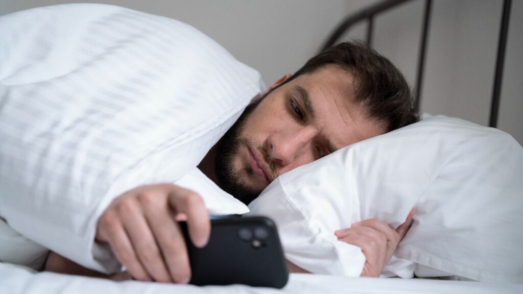  Hombre tumbado en la cama con el móvil sin poder conciliar el sueño (Fuente: Freepik)