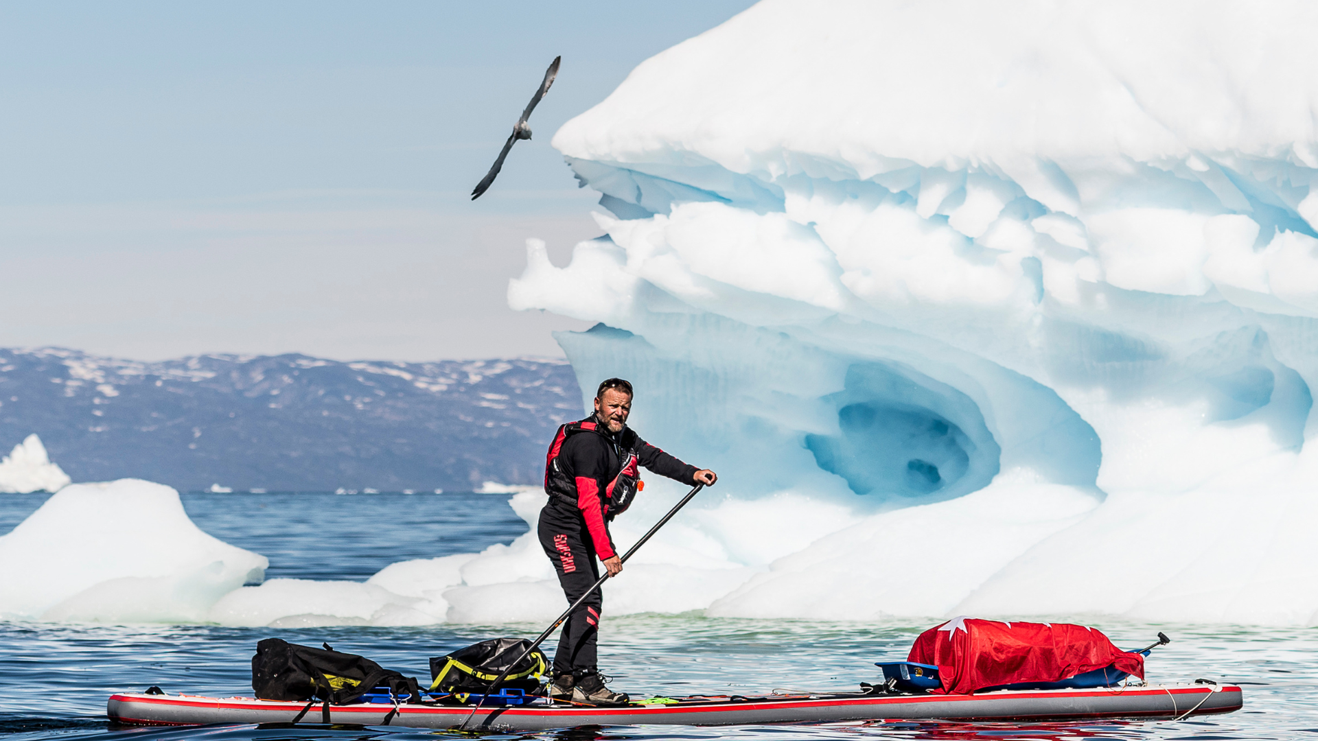 De la Rosa rema sobre su tabla de surf con un enorme bloque de hielo de fondo