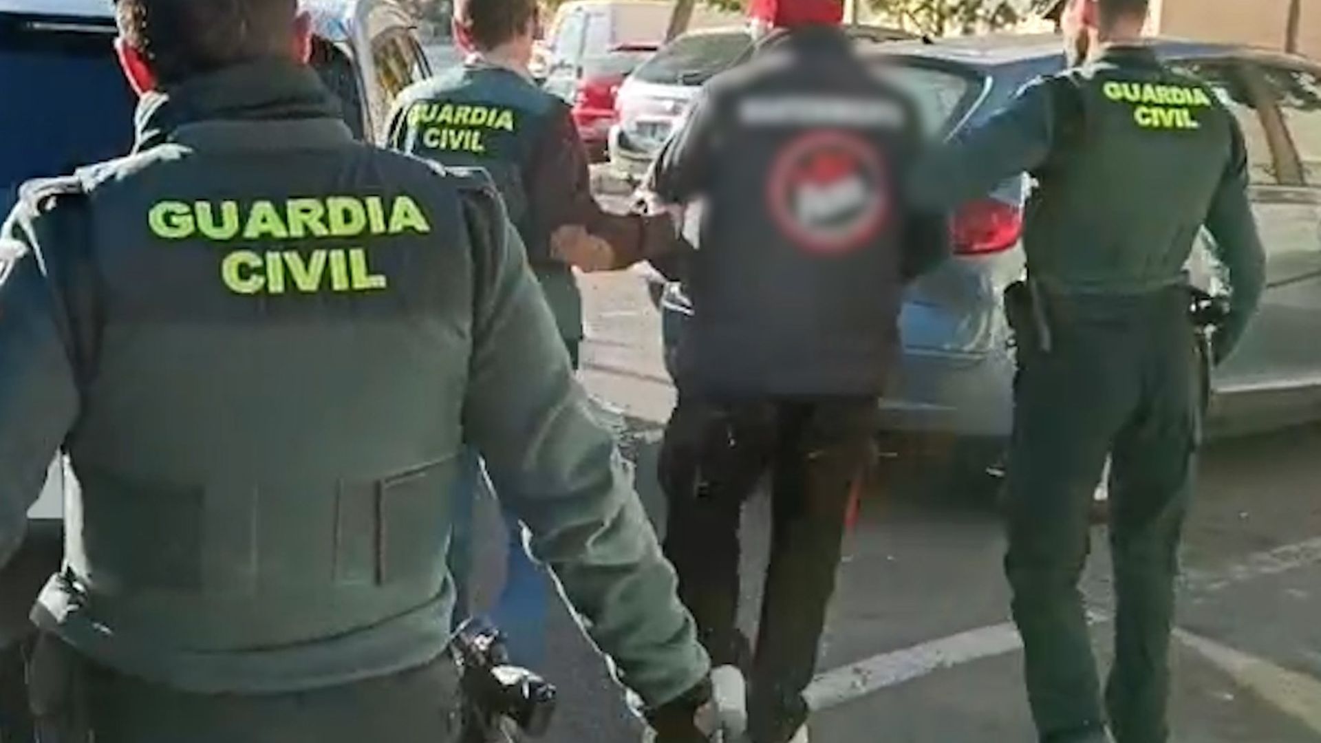 Agentes custodian al hombre arrestado, con chaqueta y gorro rojo